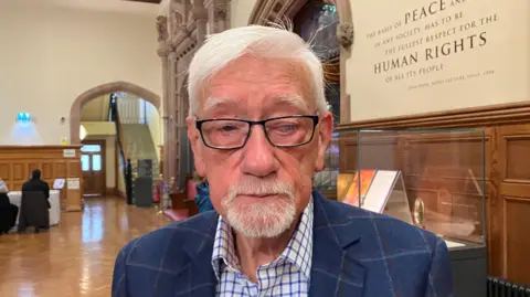 Terry Wright has short white hair and is wearing black-rimmed glasses. He is wearing a navy blue pin striped suit and a white and blue chequered suit. 