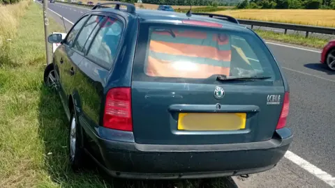 A navy blue Skoda Octavia parked on a grassy bank on the edge of the A303 on a sunny day. Half of the car is protruding out into the road. The yellow number plate has been blurred out.