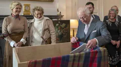 PA Media King Charles, wearing a grey suit with a white shirt and light tie, on the right of the image placing a white letter into a large wooden box with a tartan blanket partially covering it. It is taking place indoors. Sir Rod Stewart, wearing a white turtleneck jumper under a beige jacket and with a beige strap covering his torso, and wife Penny Lancaster, wearing a beige dress, watch on.