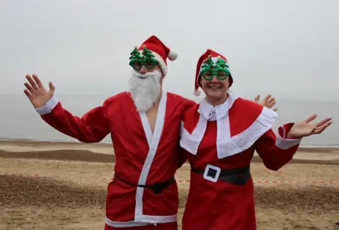 St Elizabeth Hospice Two people dressed in Father Christmas outfits on Felixstowe beach. One is wearing a beard and the other is smiling at the camera