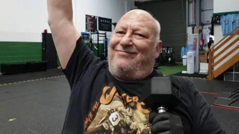 A bald man with short grey stubble is smiling into the camera as one arm is lifted and the other is holding a black weight. He is wearing a black top with a picture on it and behind him is the empty floor of a gym.
