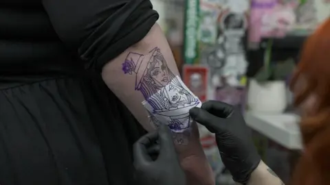 The image shows the hand of a tattoo artist wearing black gloves, peeling off the stencil of a tattoo of a woman with a hat. The arm of the person receiving the tattoo is partially covered by a black top that looks to be a black dress.