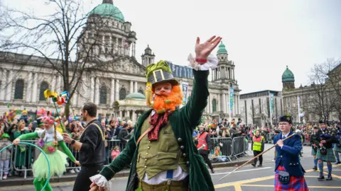 Pacemaker St Patrick's Day parade passing Belfast City Hall. At the forefront there is a man dressed as a leprechaun with bright orange beard, wearing a tall hat, long green jacket, greet waistcoat, and long red tie. He is smoking a pipe with his hand waving at the crowds. People are marching behind him playing bagpipes.