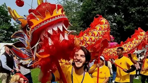 Luke McFarlane A man, Luke McFarlane at Mela festival pictured with a Chinese dragon. He is smiling, has long brunette hair and is wearing a yellow t-shirt. Behind him are other performers and trees.