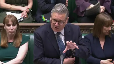 PA Media Sir Keir Starmer in a blue suit and white shirt speaking during Prime Minister's Questions in the House of Commons with colleagues seated to either side of him.