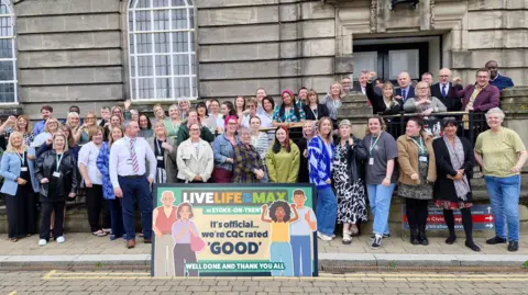 Stoke-on-Trent City Council A group of people standing outside a large council building behind a colourful sign that says "live life to the max in Stoke-on-Trent, it's official, we're CQC rated 'good', well done and thank you all". There are cartoon images of four people on the sign.
