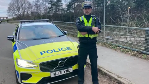 A police officer wearing a high viz vest standing in front of a police car