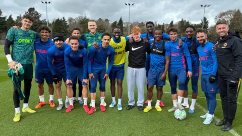 Wolverhampton Wanders football squad wearing blue training football kits in a huddle with head coach at the end and teen winner Kenzie in the middle. 