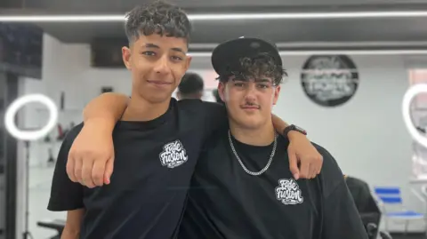 Two boys are wearing black t-shirts and have their arms over each others shoulders. They are standing in the barber shop with white ring lights in the background.