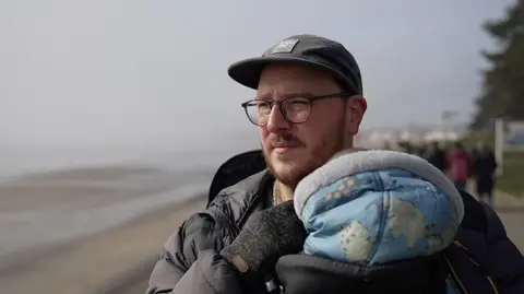 The BBC looks out of the sea in a hat and glasses. He is holding a child in a carrier