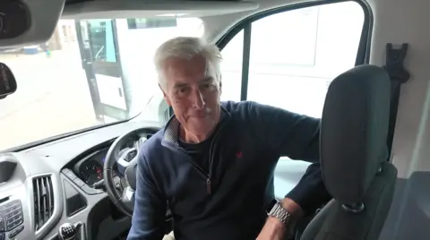 A man smiling at the camera sat in the drivers seat of a mini bus and smiling.