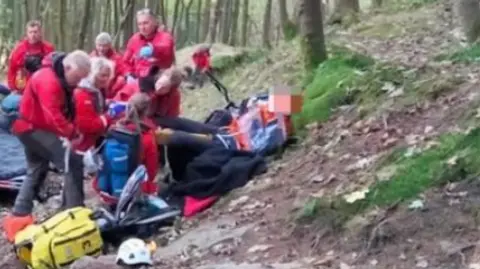 Edale Mountain Rescue Team Seven mountain rescue volunteers, in red outdoor coats, tend to a mountain bike rider who is lying on a bank in a wooded area. The rider's face is blurred.