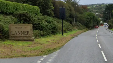 A large granite block with "Welcome to Lanner" carved into it that is sitting beside a road winding down into the village. 