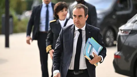 Sébastien Lecornu walks with a folder and files through a car park while wearing a dark suit.