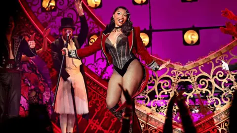Megan is wearing a red cropped jacket, a black sparkly leotard with silver stripes. She is wearing fishnet tights and black knee high boots. She is holding a black cane and smiling a the crowd. She is on stage, behind her is gold and red decorations. To her left is other members of the cast, a man in a suit and top had and a man in all black. 