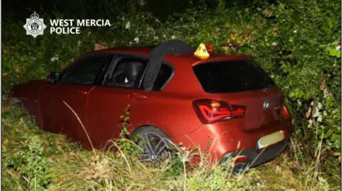 West Mercia Police A red BMW series 1 with number plate obscured in a grassy verge. It has a small yellow cone on top of it, numbered 11.