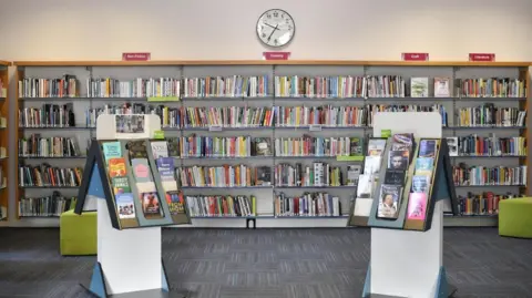 The inside of a library. There are two small book cases in the foreground, more like a sales stand in that they are triangular and allow books to be shown with the covers facing out. A large wall-to-wall shelf unit stands behind them, all full of books and with red signs above denoting subject areas. There are two green pouffes in the room, which has white walls and grey patterned carpet.