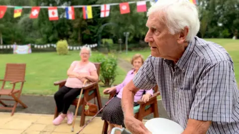 A man prepares to throw a paper plate. He is in a garden with flag bunting behind him and two other residents