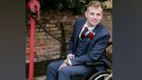 Liam in his wheelchair wearing a wedding style petrol blue suit - with a red rose on the lapel. He looks at the camera and smiles. He is on the pavement next to a brick wall.