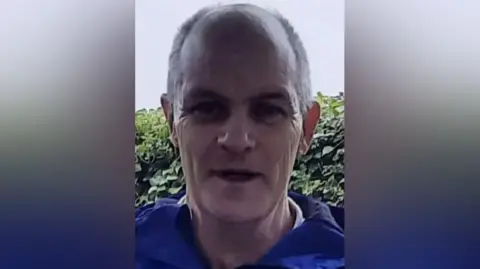 Family handout Thomas Tierney looks at the camera. He is bald with grey hair on the sides and is clean shaven. He is wearing a blue jacket with a green hedge behind him