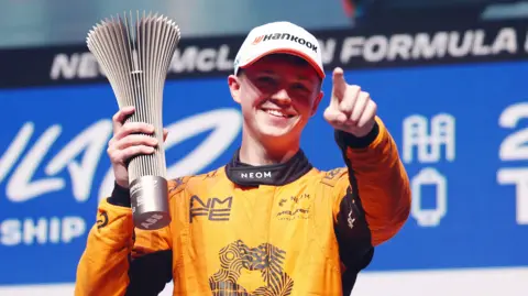 Getty Images Taylor Barnard wears an orange racing jumpsuit. His hand is outstretched and finger pointing out. In his other hand he is holding a silver trophy. 