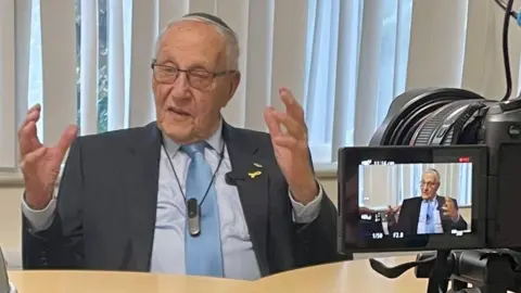 Manfred Goldberg gives a talk to 300 children in Shropshire. He is sitting at a table with a camera in front of him and gesturing with his hands as he talks about his experiences.