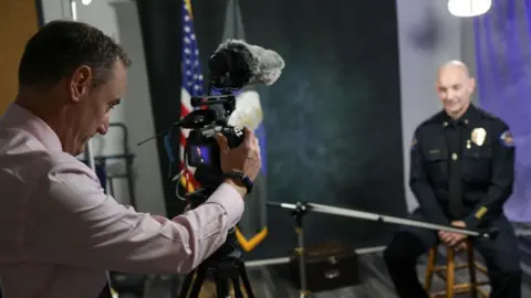 Aurora Police Department An Aurora police officer in uniform sits on a stool in a studio as a man in a pastel work shirt adjusts the camera filming him, an American flag visible in the background