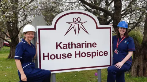 Two women in blue nurse uniforms wear hard hats next to a Katharine House Hospice.