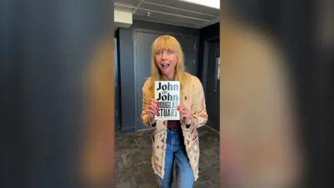 Sara Cox has blonde hair and is holding up a white book with black writing on the cover