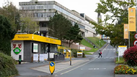 A large old university building is pictured by a long access road. To the left is a reception building with a yellow roof and the university crest.
