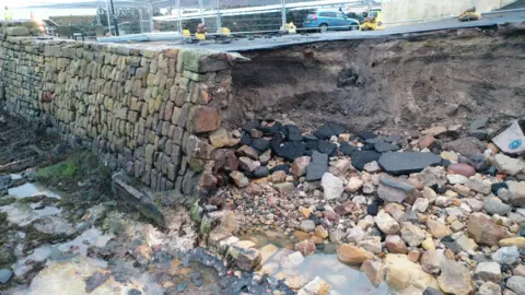 A section of broken pier - rocks and debris lying in the ground beside water - beside the rest of the cobbled pier which is standing.