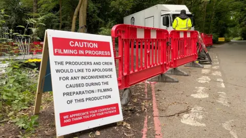 Laura Foster/BBC A white sign with a red banner and white writing is propped up on an A board, and stands next to a red plastic fence. A security guard wearing a high vis jacket stands behind the fence in front of a trailer. It is all at the edge of a road surrounded by trees. The sign reads: 'Caution. Filming in progress. The producers and crew would like to apologise for any inconvenience caused during the preparation/filming of this production. We appreciate your cooperation and thank you for your patience'.