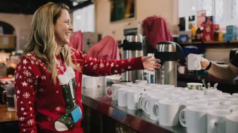 Caring at Christmas A photo showing a woman dressed in a Christmas jumper reaching for a mug. She is stood ion front of a table full of mugs and hot water containers