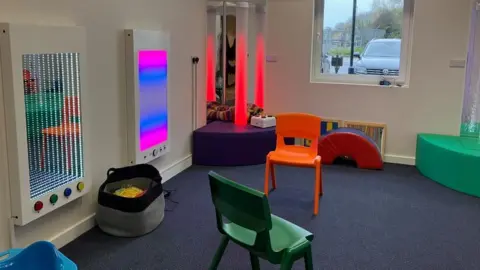 One of the sensory rooms at the new centre - a white walled room, with blue carpet and soft play furniture. On the walls are LED screens with different lighting.