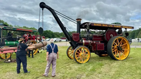 A steam engine with a rudimentary crane lifts a log, which is being guided by a man wearing a hat with a further steam engine and a couple of other people in the background.