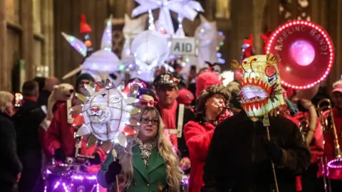 A group of people is walking along a road holding lit up lanterns of all shapes. The front two look like animal faces. Behind them are a marching brass band wearing red.
