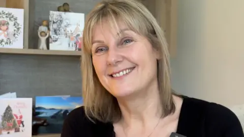 Diane Taylor has shoulder length straight blonde hair and a fringe. She is smiling, wearing a black top, and sitting in front of shelves filled with Christmas cards.