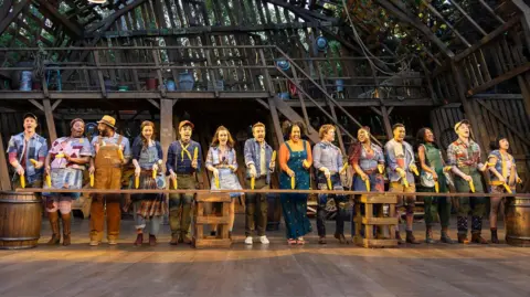 Pamela Raith The cast of Shucked the musical performing at Regent's Park Open Air Theatre, an outdoor theatre in London. A cast of 14 are on stage at the same time with corn cobs in their hands. Georgina Onuorah is fifth from the right in the row of people.