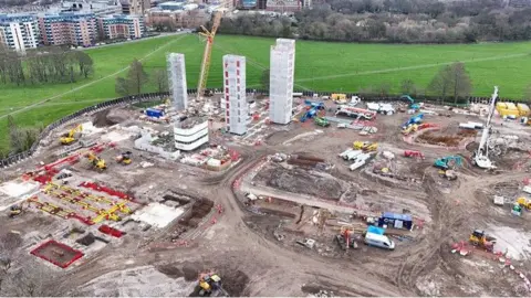 An aerial shot of building work for the Castle Leazes accommodation. The large site shows three core units of the building being erected with groundworks happening around the three shells, with lots of machinery and materials in the area. Outside the building site is a green area with more high rise buildings in the background.
