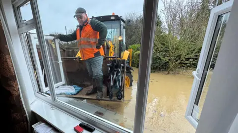 A view from inside a bungalow, looking through an open window, as a yellow digger drives through brown flood water towards it. A man wearing an orange hi-vis vest is standing in the bucket of the digger.