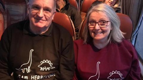 Ann Pearce wearing a red jumper and glasses sitting next to her husband Graham, on the bus.