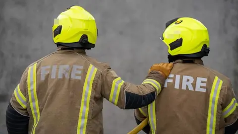 Two firefighters in mainly brown uniform, seen from the back. They are wearing yellow helmets and the firefighter on the left has a hand on a shoulder of the firefighter on the right.