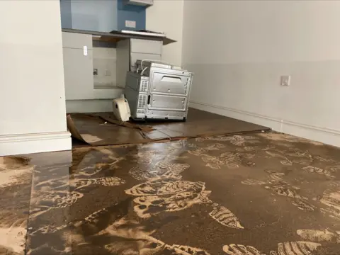 A room which shows a muddy, wet floor with footprints. There is a line about waist-high on the walls which shows how high the water went. In the background there is a kitchen applicance. 