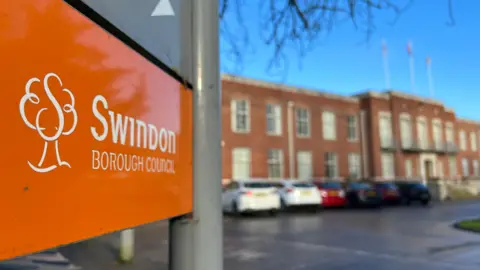 Close-up of an orange Swindon Borough Council sign, in front of a blurred red brick building which is it's headquarters.