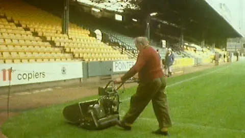 A man mowing a lawn at Norwich City Football Club. He is wearing a red top and dark trousers. You can see a badly damaged football stand, with one or two other people standing by it. 