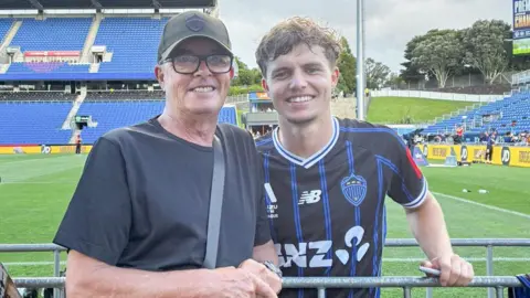 A man in a baseball cap with glasses on in a black T-Shirt stands next to a young footballer with brown hair in a black and blue striped top. They are standing in front of a football pitch with a large blue stand in the background.