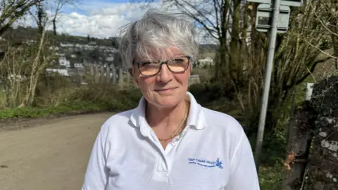 A woman with short white hair is smiling at the camera. She is wearing a t-shirt that says Visit Tamar Valley and underneath Bridging Devon and Cornwall. You can just see the viaduct over the River Tamar in the background. 