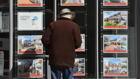 Getty Images An elderly man looks at signs for renting and buying houses outside an estate agent's shop. He's wearing a hat and a brown coat.