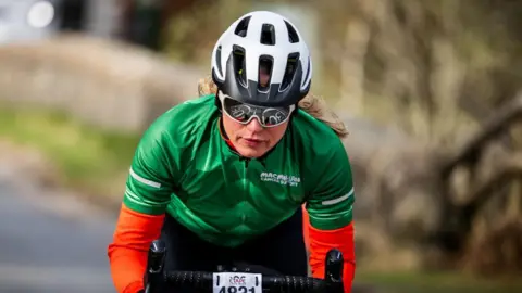 Paul Campbell A close up of a female cyclist. She is wearing cycling glasses, a black and white helmet and a green and orange cycling top.