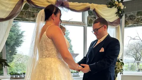 Contributed Lauren and Jack are pictured during their marriage blessing ceremony. They are facing each other and holding hands. Lauren is in her white wedding dress with a long veil placed behind her head. Jack is wearing a navy suit with a white shirt. He has short dark hair and is wearing black glasses.